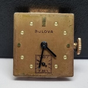 Puede incluir: Un movimiento de reloj Bulova vintage con una esfera cuadrada de color dorado. La esfera del reloj presenta agujas negras, un peque&ntilde;o dial de segundos y el nombre de la marca Bulova. El reloj tiene una corona dorada y peque&ntilde;os detalles dorados.