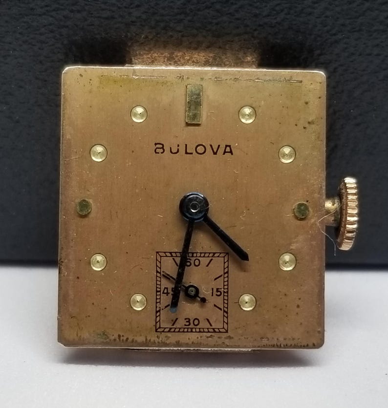 Puede incluir: Un movimiento de reloj Bulova vintage con una esfera cuadrada de color dorado. La esfera del reloj presenta agujas negras, un peque&ntilde;o dial de segundos y el nombre de la marca Bulova. El reloj tiene una corona dorada y peque&ntilde;os detalles dorados.
