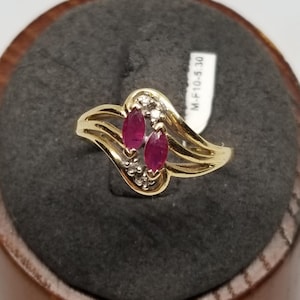 May include: A gold ring featuring two marquise-cut, deep red gemstones and small accent diamonds. The ring has a flowing, asymmetrical design. A tag with the text "M-F10-5 30" is attached.