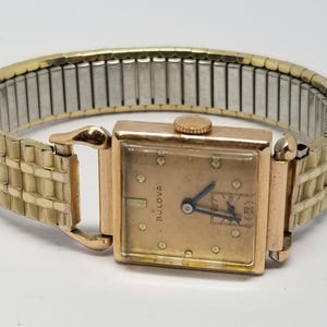 Puede incluir: Un reloj de pulsera Bulova vintage con una esfera rectangular dorada y una correa extensible dorada. La esfera del reloj es de color oro claro con agujas negras y peque&ntilde;os marcadores. La correa es una mezcla de metal dorado y plateado.