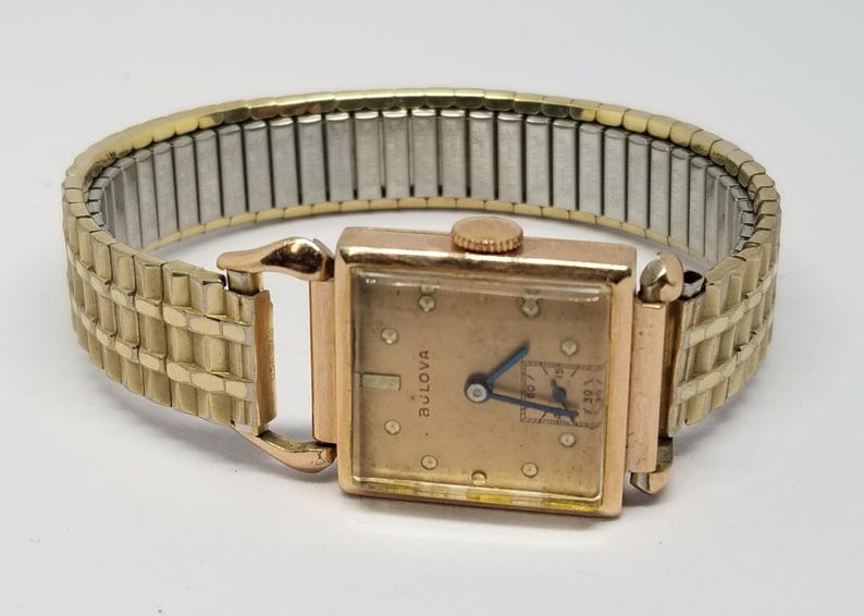 Puede incluir: Un reloj de pulsera Bulova vintage con una esfera rectangular dorada y una correa extensible dorada. La esfera del reloj es de color oro claro con agujas negras y peque&ntilde;os marcadores. La correa es una mezcla de metal dorado y plateado.
