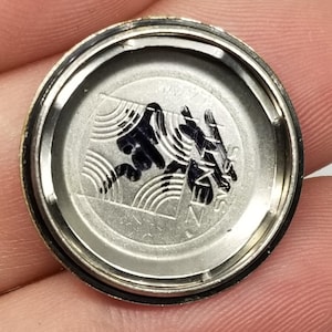 May include: A silver-toned watch back with a black graphic design. The design includes stylized waves and text. The watch back is circular with a dark rim and is held by a person's fingers.