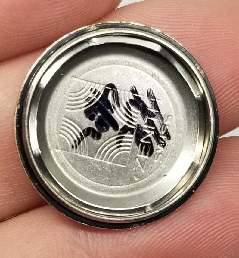 May include: A silver-toned watch back with a black graphic design. The design includes stylized waves and text. The watch back is circular with a dark rim and is held by a person's fingers.
