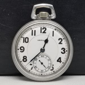 May include: A silver-toned pocket watch with a white face and black numerals. The watch features a smaller seconds dial and the brand name "HAMILTON". The watch has a decorative loop at the top for a chain.