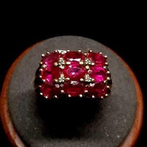 May include: A gold-toned ring featuring a cluster of oval-cut, vibrant red gemstones. Small accent stones are interspersed among the larger gems. The ring is set on a dark, textured surface.