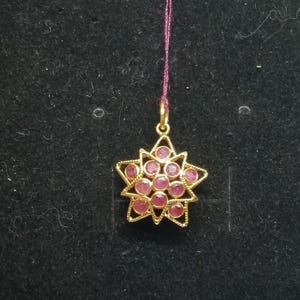 May include: A gold-colored pendant in the shape of a star, featuring a cluster of pink gemstones. The pendant is suspended from a thin, purple string. The star design has a detailed, openwork pattern.