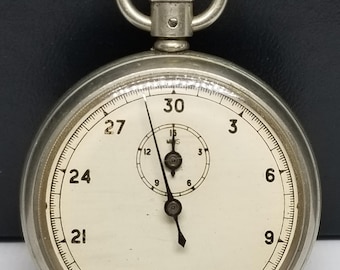 Puede incluir: Un cronómetro vintage con caja plateada y esfera blanca. El reloj presenta números y marcas en negro, con una esfera más pequeña en la parte superior. Las manecillas señalan 12 segundos y 0 segundos. Tiene una corona de cuerda y un anillo en la parte superior.