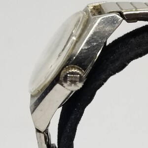 May include: A vintage silver-toned wristwatch with a clear face and a metal band. The watch is shown from a side angle, highlighting the crown and the details of the band's links. The watch is resting on a black surface.