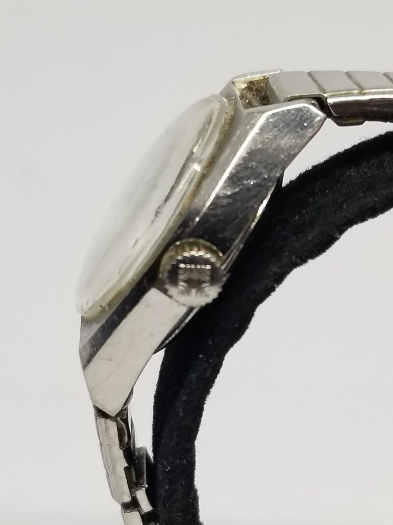 May include: A vintage silver-toned wristwatch with a clear face and a metal band. The watch is shown from a side angle, highlighting the crown and the details of the band's links. The watch is resting on a black surface.