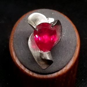 May include: A silver ring featuring a large, pear-shaped, vibrant red gemstone. The gemstone is set in a silver mounting with stylized, curved accents. The ring is displayed on a dark, textured surface.