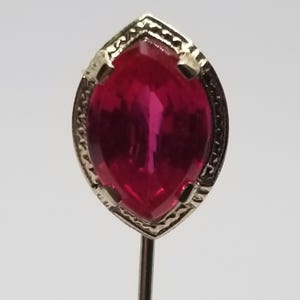 May include: A vintage-style pin with a marquise-cut, deep red gemstone set in a silver-toned metal frame. The frame has a decorative, textured border. The pin is on a thin, silver-colored stem.