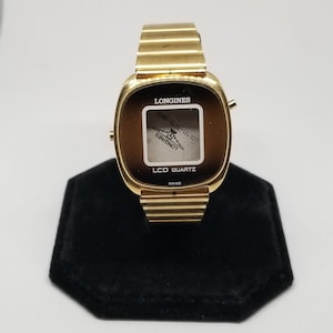 May include: A gold-toned Longines LCD Quartz watch with a square face and a brown digital display. The watch has a gold-toned metal band.