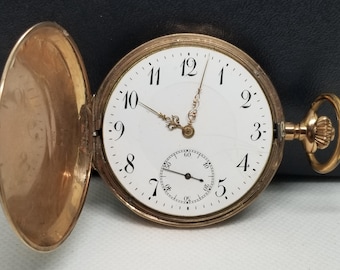 Puede incluir: Un reloj de bolsillo dorado con esfera blanca y números negros. El reloj tiene una subesfera y agujas ornamentadas. La tapa está abierta, revelando el mecanismo interno. El reloj está sobre un fondo oscuro.