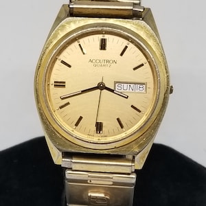 May include: A gold-toned Accutron Quartz wristwatch with a gold-tone metal band. The watch face is gold with black hour markers and hands. The date window displays "SUN 18".