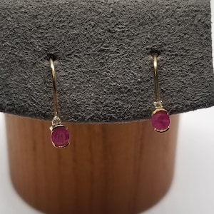 May include: A pair of gold drop earrings featuring a single round, deep pinkish-red gemstone at the base of each curved wire. Each earring includes a small, clear accent stone positioned directly above the primary colored gemstone setting.