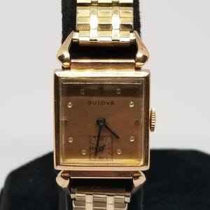 Puede incluir: Un reloj de pulsera Bulova vintage con una esfera rectangular dorada y una correa dorada a juego. La esfera del reloj muestra la marca Bulova y una peque&ntilde;a esfera secundaria. El reloj se exhibe sobre un soporte negro.