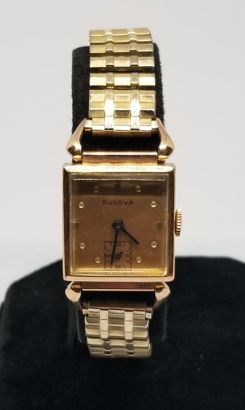 Puede incluir: Un reloj de pulsera Bulova vintage con una esfera rectangular dorada y una correa dorada a juego. La esfera del reloj muestra la marca Bulova y una peque&ntilde;a esfera secundaria. El reloj se exhibe sobre un soporte negro.
