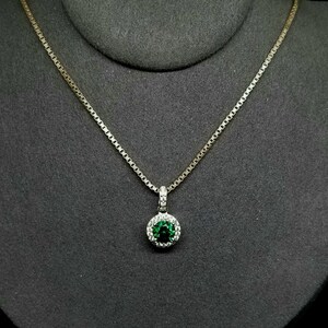 May include: A gold-toned necklace with a round, emerald-colored pendant. The pendant is surrounded by a halo of small, clear stones. The necklace is displayed on a dark gray surface.