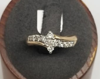 May include: A gold ring with a cluster of small, clear stones in the center and smaller stones along the band. The ring is set against a dark gray background and a wooden stand.