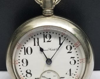 Puede incluir: Reloj de bolsillo plateado con esfera blanca, manecillas negras y números negros. La esfera del reloj incluye una esfera secundaria más pequeña y el texto "Illinois Watch Co." El reloj tiene una corona texturizada y un lazo para una cadena.