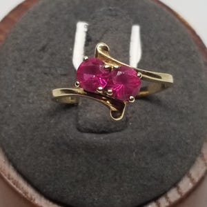 May include: A gold ring featuring two oval-cut, vibrant pink gemstones. The ring has a swirling design with the gemstones set side-by-side. The ring is displayed on a dark gray surface.
