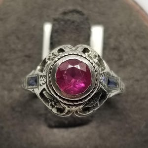 May include: A silver ring featuring a large, round, ruby-colored gemstone at the center. The ring has intricate floral and filigree detailing, with two smaller blue gemstones on either side of the main stone. The ring is set against a neutral background.