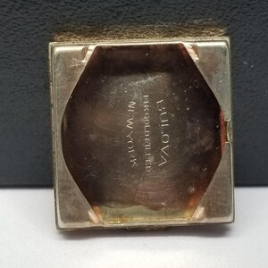 Puede incluir: Primer plano de la parte trasera de un reloj Bulova vintage. La caja del reloj es dorada y rectangular con un interior octogonal. El texto "BULOVA 14K GOLD FILLED NEW YORK" es visible en el interior del reloj.