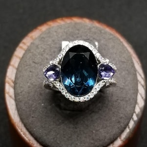 May include: A silver ring featuring a large, oval-cut, dark blue gemstone as the centerpiece, flanked by two smaller, pear-shaped, purple gemstones. The ring is accented with small, clear crystals around the main stone and along the band.