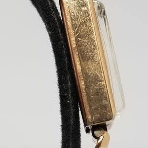 Puede incluir: Un reloj de pulsera rectangular vintage en tono dorado con una esfera transparente. El reloj est&aacute; sujeto a una correa negra. La caja del reloj y los conectores de la correa son dorados. El reloj es de estilo cl&aacute;sico.