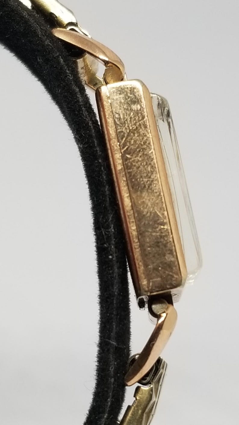 Puede incluir: Un reloj de pulsera rectangular vintage en tono dorado con una esfera transparente. El reloj est&aacute; sujeto a una correa negra. La caja del reloj y los conectores de la correa son dorados. El reloj es de estilo cl&aacute;sico.