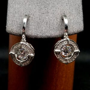 May include: A pair of silver-toned hoop earrings. Each earring features a central, clear, round gemstone surrounded by smaller, clear gemstones. The design includes a circular frame with a cross-like element.