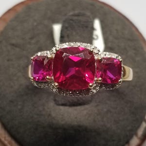 May include: A gold ring featuring three large, vibrant pink gemstones. The central stone is square-cut, flanked by two smaller, square-cut stones. Each stone is surrounded by a halo of small, clear accent stones, creating a sparkling effect.
