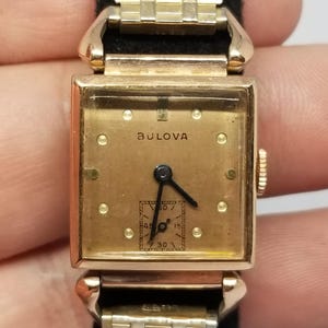 Puede incluir: Un reloj de pulsera Bulova vintage con una esfera cuadrada dorada y una correa dorada a juego. La esfera del reloj muestra el nombre de la marca Bulova y peque&ntilde;os marcadores circulares. El reloj tiene agujas negras y un subdial para los segundos.