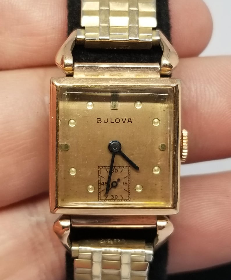 Puede incluir: Un reloj de pulsera Bulova vintage con una esfera cuadrada dorada y una correa dorada a juego. La esfera del reloj muestra el nombre de la marca Bulova y peque&ntilde;os marcadores circulares. El reloj tiene agujas negras y un subdial para los segundos.