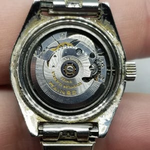 May include: A silver-toned Zenith watch with the back removed, revealing the intricate inner workings of the watch mechanism. The watch face is visible, with the words "Zenith" and "Swiss" clearly displayed. The watch band is also silver.