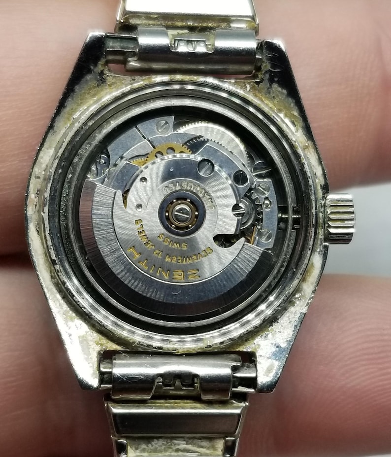May include: A silver-toned Zenith watch with the back removed, revealing the intricate inner workings of the watch mechanism. The watch face is visible, with the words "Zenith" and "Swiss" clearly displayed. The watch band is also silver.