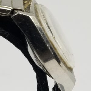 May include: Close-up of a vintage silver-toned wristwatch. The watch has a rectangular face with a clear crystal and a silver band. The watch is resting on a black surface.