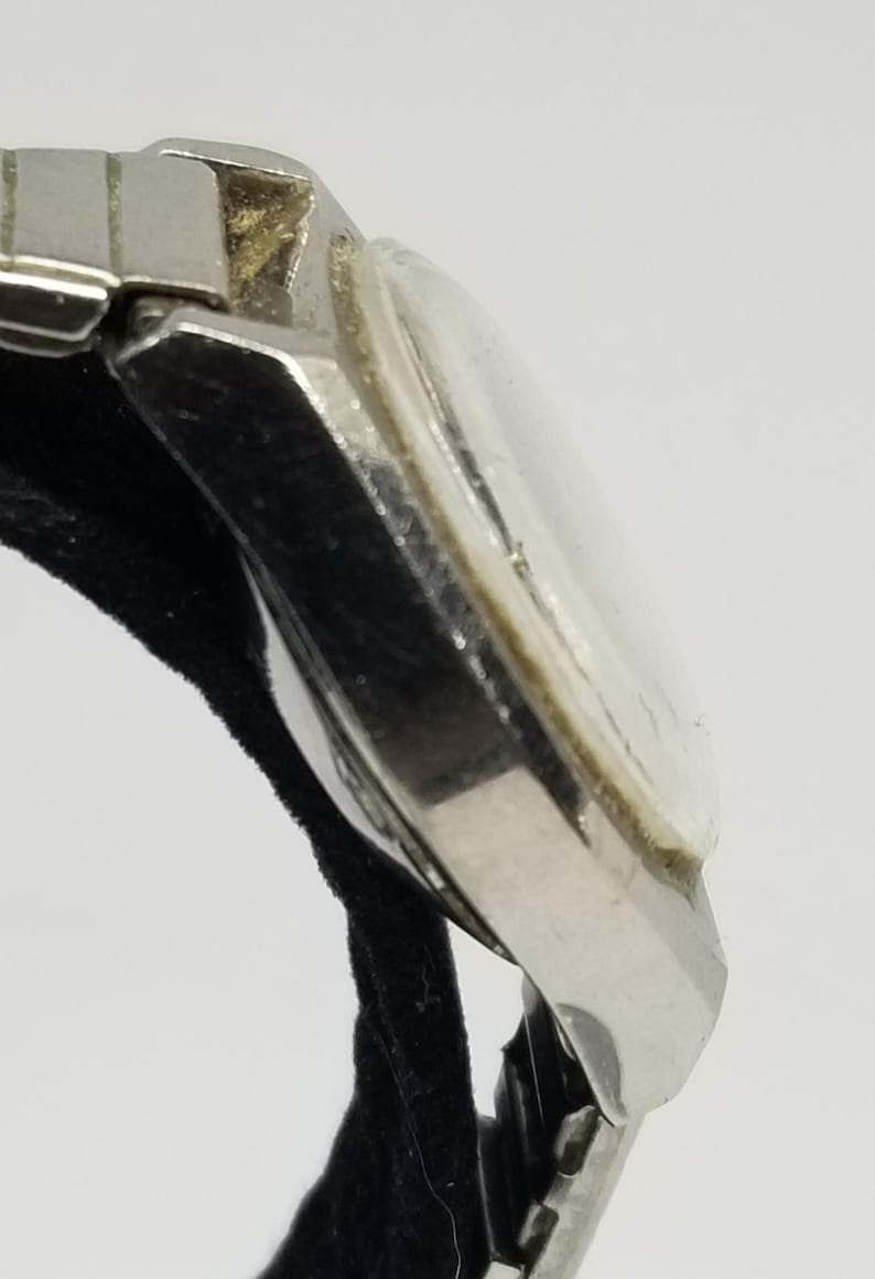 May include: Close-up of a vintage silver-toned wristwatch. The watch has a rectangular face with a clear crystal and a silver band. The watch is resting on a black surface.