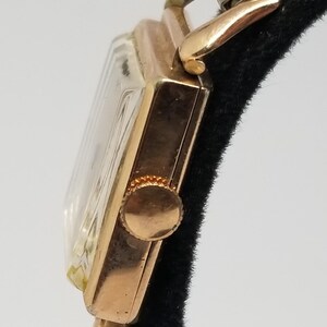 Puede incluir: Primer plano de un reloj de pulsera vintage de oro. El reloj tiene una esfera rectangular con un cristal transparente y una correa dorada. La corona del reloj es visible en el lateral. El reloj se muestra sobre un fondo negro.