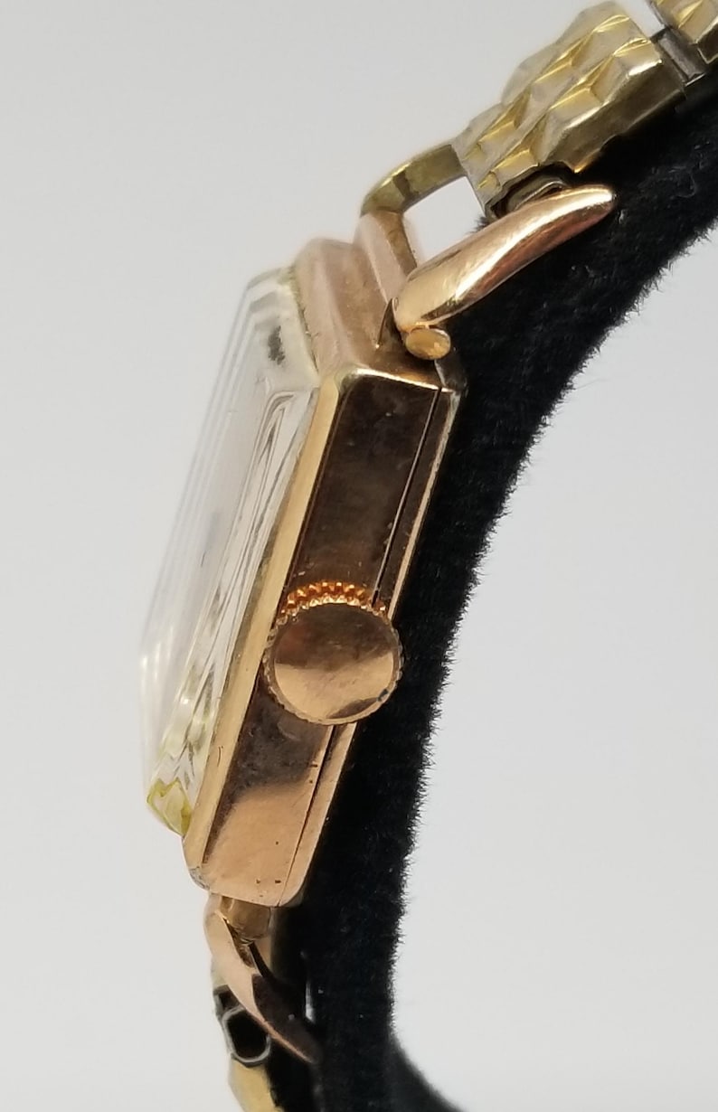 Puede incluir: Primer plano de un reloj de pulsera vintage de oro. El reloj tiene una esfera rectangular con un cristal transparente y una correa dorada. La corona del reloj es visible en el lateral. El reloj se muestra sobre un fondo negro.