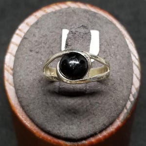 May include: A silver-toned ring featuring a smooth, round, polished black stone set in the center. The band consists of two thin, curved metal segments that wrap around the stone, creating an open, eye-like design on both sides of the setting.