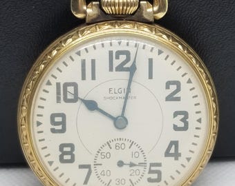 Puede incluir: Un reloj de bolsillo Elgin Shockmaster dorado con esfera blanca y números negros. El reloj tiene un pequeño dial de segundos y un lazo dorado para una cadena. Las manecillas del reloj son azules.