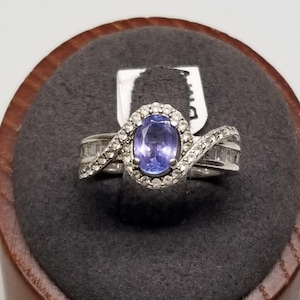 May include: A silver ring with a purple oval gemstone surrounded by smaller white gemstones. The ring has a twisted band with additional white gemstones.