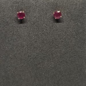 May include: A pair of stud earrings featuring round, deep red gemstones set in gold-colored metal. The earrings are displayed on a dark gray textured surface, showcasing their color and design.