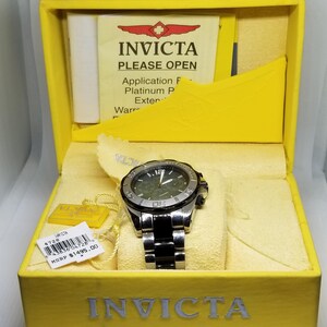 May include: An Invicta watch in a yellow box. The watch has a silver and black band and a dark face. The box has the Invicta logo and the text "PLEASE OPEN". A price tag is visible.