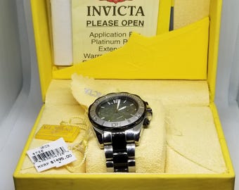 May include: An Invicta watch in a yellow box. The watch has a silver and black band and a dark face. The box has the Invicta logo and the text "PLEASE OPEN". A price tag is visible.