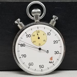 May include: A vintage stopwatch with a white face and black numerals. The stopwatch has a silver metal casing, two side buttons, and a top ring. The face includes a smaller inner dial and a second hand. The time is approximately 15 seconds.