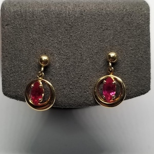 May include: A pair of gold-colored earrings with a dangling design. Each earring features a small, oval-shaped, red gemstone set within a gold circle. The earrings are displayed on a gray surface.