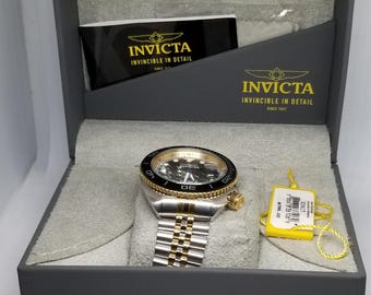 May include: A silver and gold Invicta watch with a black dial, presented in a gray box. The watch features a two-tone metal band and a gold-colored bezel. The box has the Invicta logo and the words "Invincible in Detail."