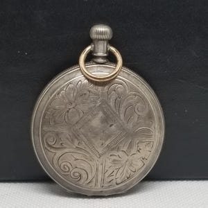 May include: A silver pocket watch with intricate floral and scroll engravings. The watch features a gold-colored ring and a textured crown for winding. The face has a diamond-shaped design in the center.